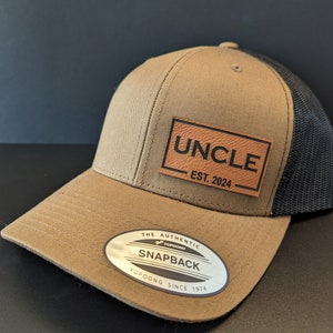 Könnte beinhalten: Eine braune und schwarze Baseballkappe mit einem braunen Lederpatch, auf dem "UNCLE EST. 2024" steht. Die Kappe hat ein schwarzes Netzrücken und ein silbernes YUPOONG-Etikett mit der Aufschrift "THE AUTHENTIC SNAPBACK YUPOONG SINCE 1974".