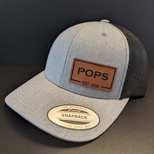 May include: A gray and black mesh baseball cap with a brown leather patch that says "POPS EST. 2024". The cap has a gold sticker that says "THE AUTHENTIC YUPOONG SNAPBACK YUPOONG SINCE 1974".