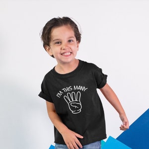 May include: A young child wearing a black t-shirt with the text "I'm this many" and a white hand with three fingers raised. The child is standing on a blue and teal background.