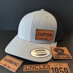 May include: A grey and black trucker cap with a brown leather patch that says "CUSTOM EST. 2024". Additional brown leather patches with the words "HUSBAND EST. 2024", "UNCLE", "DOG NAME", and "VINTAGE 1959" are also included.