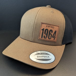 May include: A brown and black baseball cap with a brown leather patch that says "Vintage 1964". The cap has a black mesh back and a white sticker that says "The Authentic Yupoong Snapback Yupoong Since 1974".