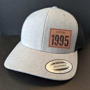 Puede incluir: Una gorra de camionero de malla gris y negra con un parche de cuero marrón que dice "Vintage 1995". La gorra tiene una parte trasera de malla negra y una parte delantera gris con una pegatina ovalada blanca que dice "The Authentic Yupoong Snapback Yupoong Since 1974".