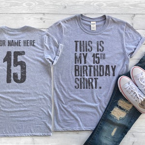May include: Gray t-shirt with the text "This is my 15th birthday shirt." on the front and the number "15" on the back. The shirt is shown with a pair of jeans and white sneakers.
