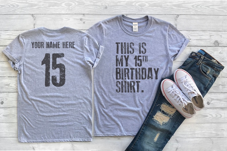 May include: Gray t-shirt with the text "This is my 15th birthday shirt." on the front and the number "15" on the back. The shirt is shown with a pair of jeans and white sneakers.