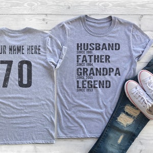 May include: Two gray t-shirts, one with the text "YOUR NAME HERE 70" and the other with the text "HUSBAND SINCE 1981 FATHER SINCE 1984 GRANDPA SINCE 2005 LEGEND SINCE 1953".