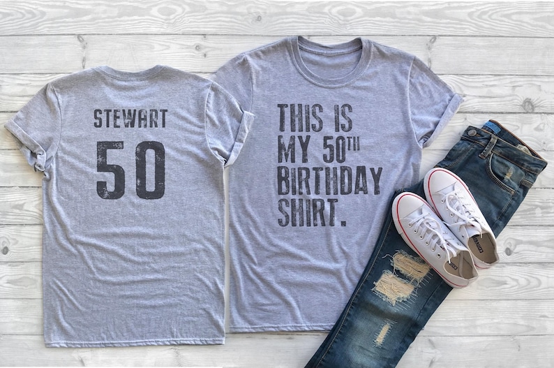May include: Two gray t-shirts. One shirt has the text "STEWART 50" on the back. The other shirt has the text "THIS IS MY 50th BIRTHDAY SHIRT." on the front.