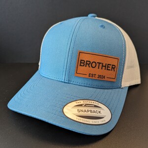 May include: A light blue and white baseball cap with a brown leather patch that says "Brother Est. 2024". The cap has a white mesh back and a black "The Authentic Yupoong Snapback Yupoong Since 1974" label.