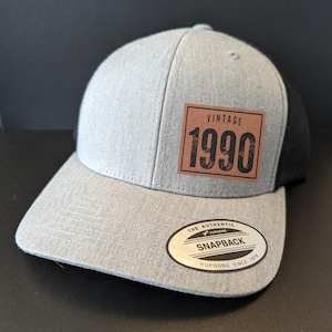 May include: Gray and black mesh trucker hat with a brown leather patch that says "Vintage 1990". The hat has a black mesh back and a gray front with a YUPOONG SNAPBACK label.