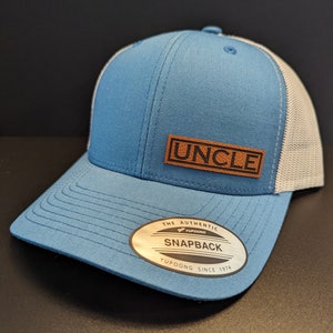 Può includere: Un cappello da baseball blu e bianco con una toppa in pelle marrone che dice "UNCLE". Il cappello ha una parte posteriore in rete e un logo YUPOONG sul lato.