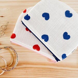 Peut inclure: Deux pochettes en tissu avec fermetures éclair. La pochette du haut est blanche avec des motifs de cœurs bleus. La pochette du bas est blanche avec des motifs de cœurs rouges. Les deux pochettes ont une fermeture éclair rose.