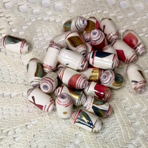 May include: A collection of cylindrical beads featuring a white base with layered patterns in red, gold, and dark blue. Each bead has a central hole and a rolled paper construction, displaying varied geometric shapes and metallic accents across the surface.
