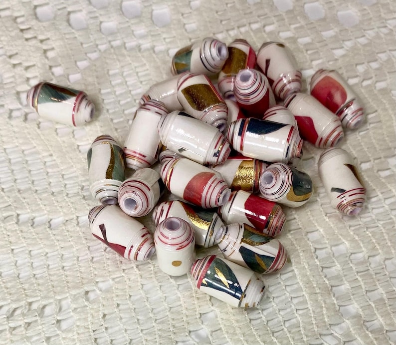 May include: A collection of cylindrical beads featuring a white base with layered patterns in red, gold, and dark blue. Each bead has a central hole and a rolled paper construction, displaying varied geometric shapes and metallic accents across the surface.