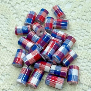 May include: A collection of small, cylindrical beads with a plaid pattern in red, white, and blue. The beads are scattered on a white, textured surface, creating a visually appealing arrangement. These beads are suitable for jewelry making and craft projects.
