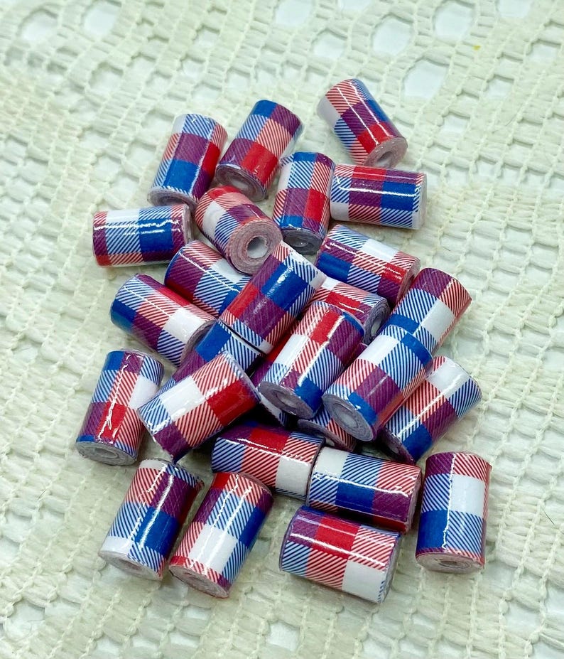 May include: A collection of small, cylindrical beads with a plaid pattern in red, white, and blue. The beads are scattered on a white, textured surface, creating a visually appealing arrangement. These beads are suitable for jewelry making and craft projects.