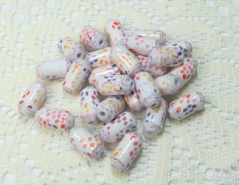 May include: A pile of white, orange and purple polka dot beads. The beads are made of paper and are rolled into a cylinder shape.