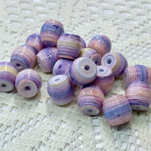 K&ouml;nnte beinhalten: Eine Ansammlung von tonnenf&ouml;rmigen Perlen mit einem geschichteten, gerollten Papierdesign. Jede Perle zeigt einen Verlauf aus sanften Pastellfarben, darunter Lavendel, Hellviolett, Pfirsich und Creme, mit einer gl&auml;nzenden Oberfl&auml;che und Loch.