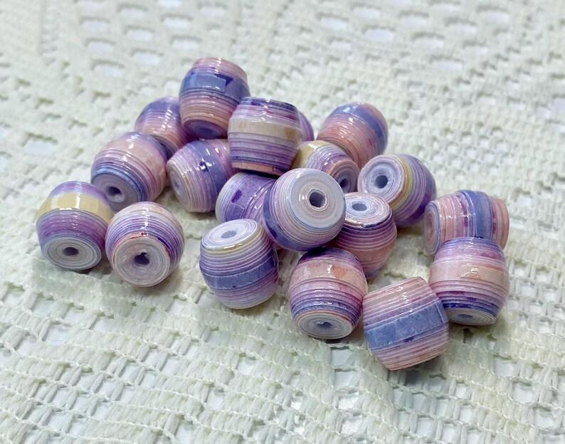 K&ouml;nnte beinhalten: Eine Ansammlung von tonnenf&ouml;rmigen Perlen mit einem geschichteten, gerollten Papierdesign. Jede Perle zeigt einen Verlauf aus sanften Pastellfarben, darunter Lavendel, Hellviolett, Pfirsich und Creme, mit einer gl&auml;nzenden Oberfl&auml;che und Loch.
