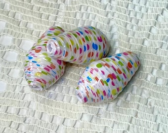 May include: Three white, oblong paper beads with colorful polka dots in shades of pink, blue, green, and yellow. The beads are arranged on a white, textured surface. The beads have a cylindrical shape with a hole running through the center.