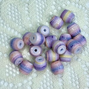 K&ouml;nnte beinhalten: Eine Ansammlung von tonnenf&ouml;rmigen Perlen mit einem geschichteten, mehrfarbigen Design in Lavendel, zartem Rosa, Creme und Dunkelviolett. Jede Perle hat eine glatte, gl&auml;nzende Oberfl&auml;che und ein Mittelloch, das eine gerollte Papierkonstruktion zeigt.