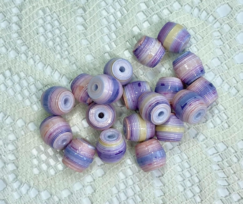 K&ouml;nnte beinhalten: Eine Ansammlung von tonnenf&ouml;rmigen Perlen mit einem geschichteten, mehrfarbigen Design in Lavendel, zartem Rosa, Creme und Dunkelviolett. Jede Perle hat eine glatte, gl&auml;nzende Oberfl&auml;che und ein Mittelloch, das eine gerollte Papierkonstruktion zeigt.