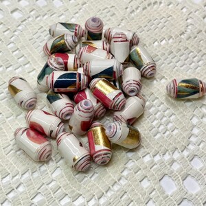 May include: A collection of cylindrical paper beads featuring a white base with abstract patterns in shades of red, dark green, and metallic gold. Each bead has a central hole and a smooth, glossy finish, arranged in a loose pile.