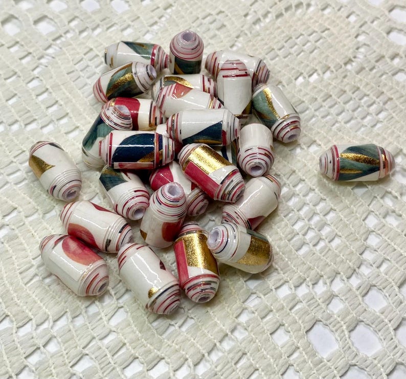 May include: A collection of cylindrical paper beads featuring a white base with abstract patterns in shades of red, dark green, and metallic gold. Each bead has a central hole and a smooth, glossy finish, arranged in a loose pile.