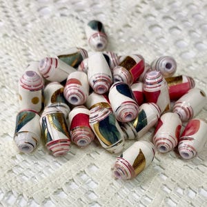 May include: A collection of cylindrical paper beads featuring a white base with assorted patterns in red, dark green, and metallic gold. Each bead has a central hole and is constructed from tightly rolled paper strips with visible layered edges.