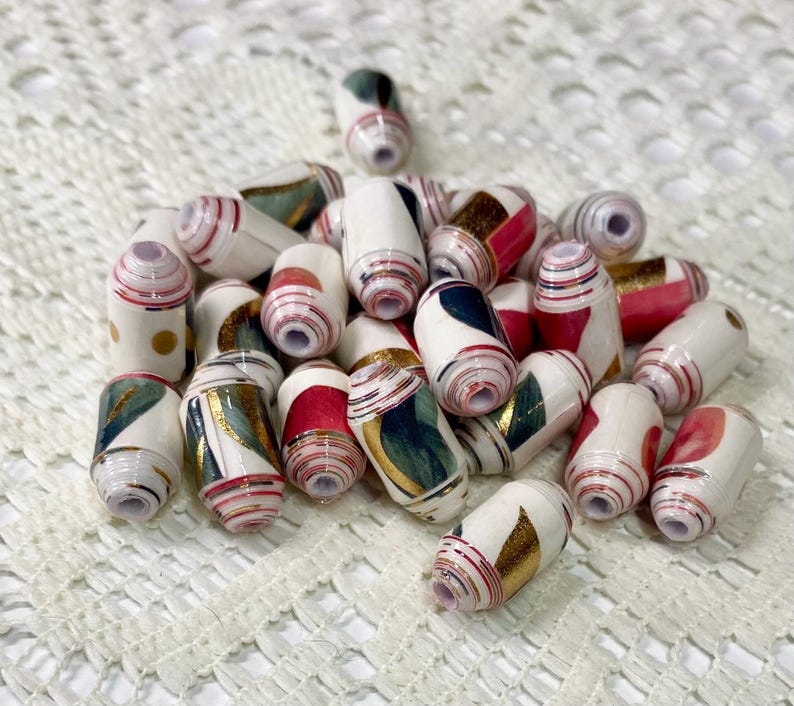 May include: A collection of cylindrical paper beads featuring a white base with assorted patterns in red, dark green, and metallic gold. Each bead has a central hole and is constructed from tightly rolled paper strips with visible layered edges.