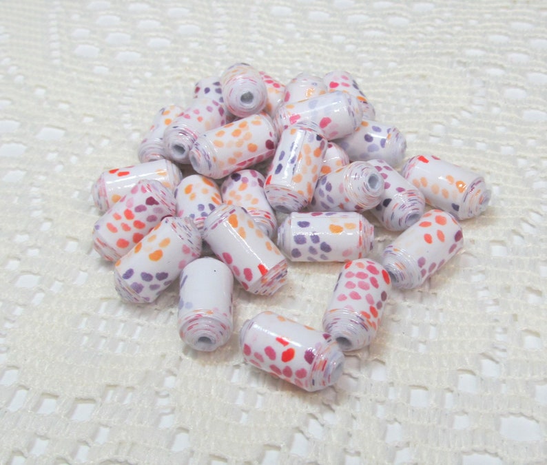 May include: A pile of white paper beads with orange, pink, and purple polka dots. The beads are arranged in a random pattern on a white crocheted surface.