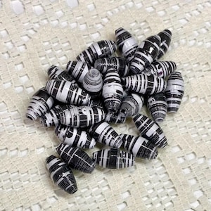 May include: A collection of barrel-shaped beads crafted from rolled paper, featuring a distinct black and white striped pattern. Each bead displays a tightly wound, layered construction with varying widths of monochromatic bands throughout the surface.