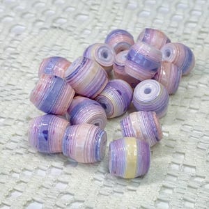 K&ouml;nnte beinhalten: Eine Ansammlung von tonnenf&ouml;rmigen Perlen aus geschichtetem, gerolltem Papier. Jede Perle zeigt ein Streifenmuster in sanften Pastellt&ouml;nen wie Rosa, Lavendel und Creme, mit einer gl&auml;nzenden Oberfl&auml;che und einem mittigen Loch zum Auff&auml;deln.