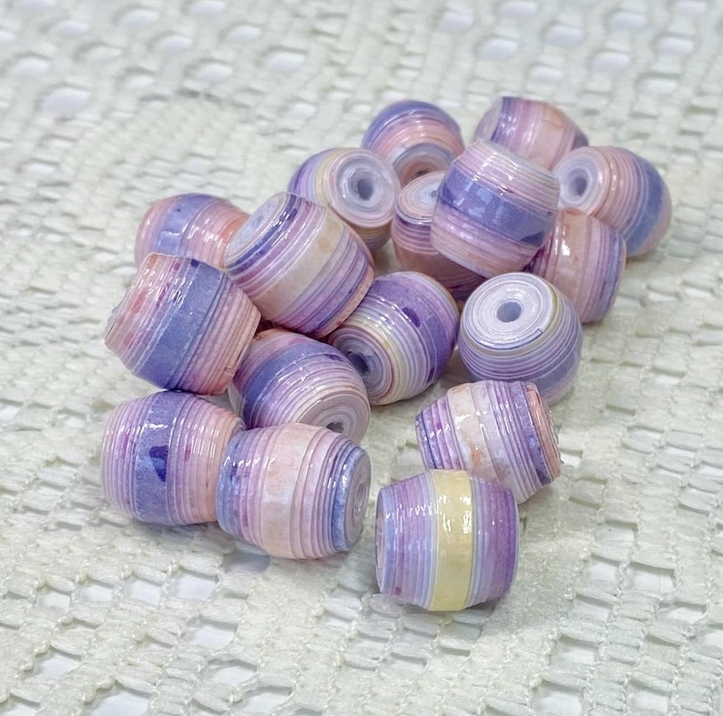 K&ouml;nnte beinhalten: Eine Ansammlung von tonnenf&ouml;rmigen Perlen aus geschichtetem, gerolltem Papier. Jede Perle zeigt ein Streifenmuster in sanften Pastellt&ouml;nen wie Rosa, Lavendel und Creme, mit einer gl&auml;nzenden Oberfl&auml;che und einem mittigen Loch zum Auff&auml;deln.