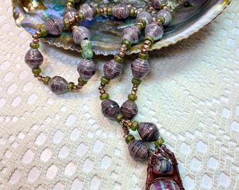 May include: A long necklace made of brown and white paper beads with green and copper accents. The necklace has a pendant made of a brown paper bead with a smaller paper bead inside.