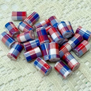 May include: A collection of small, cylindrical beads with a plaid pattern in red, white, and blue. The beads are scattered on a white, textured surface, creating a visually appealing arrangement. These beads are suitable for jewelry making.