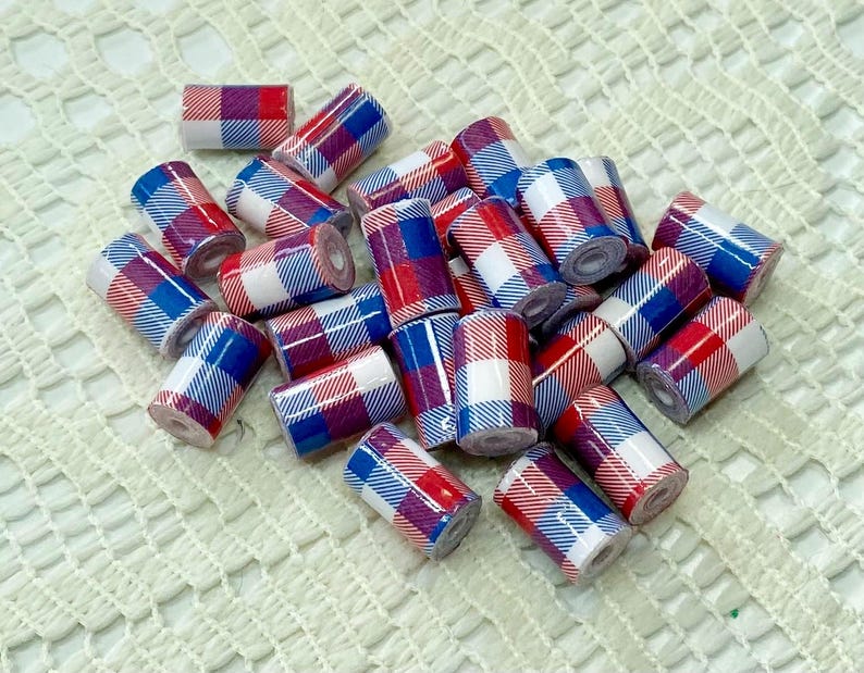 May include: A collection of small, cylindrical beads with a plaid pattern in red, white, and blue. The beads are scattered on a white, textured surface, creating a visually appealing arrangement. These beads are suitable for jewelry making.
