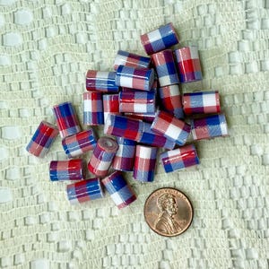 May include: A collection of small, cylindrical beads with a red, white, and blue plaid pattern. A US penny is included for size reference. The beads are scattered on a textured, off-white surface.