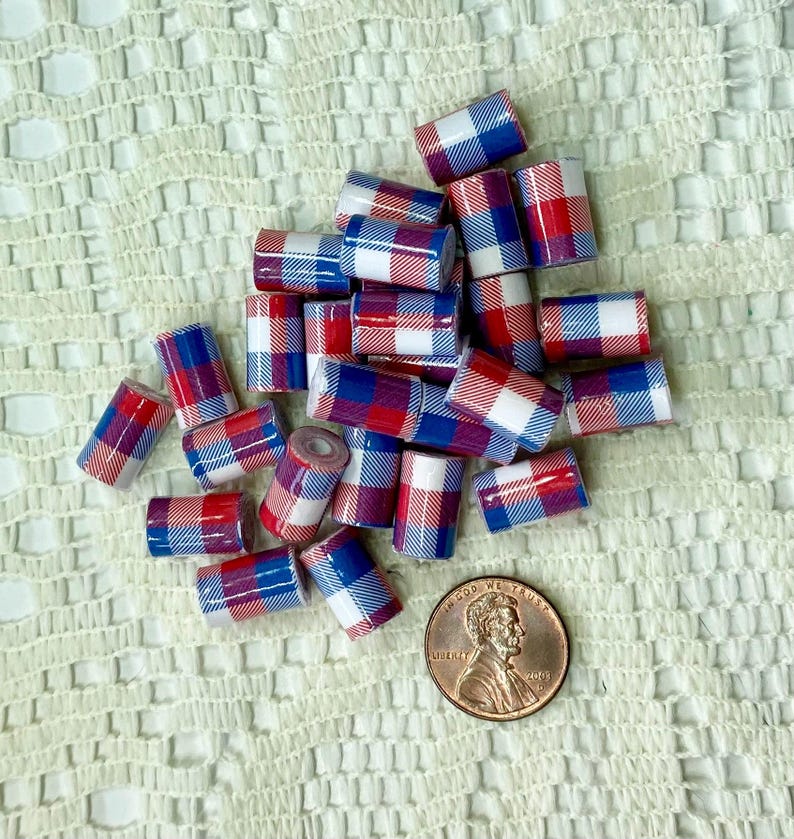 May include: A collection of small, cylindrical beads with a red, white, and blue plaid pattern. A US penny is included for size reference. The beads are scattered on a textured, off-white surface.