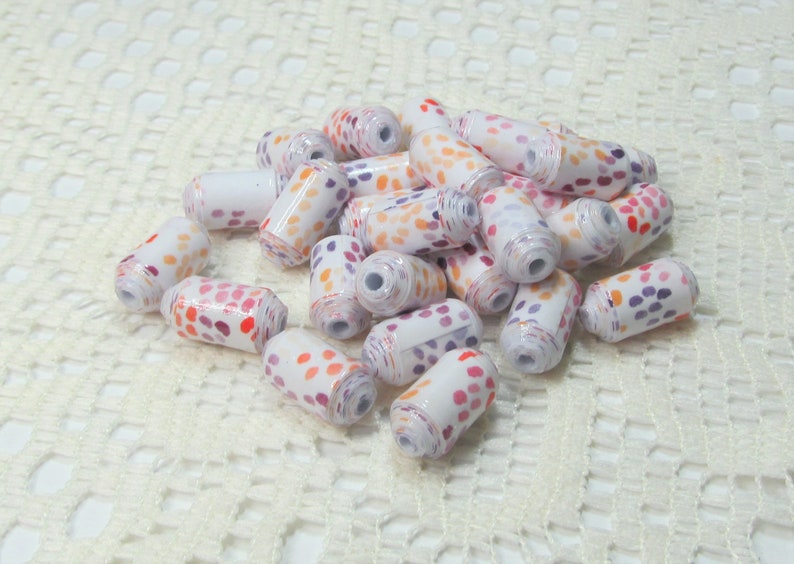 May include: A pile of white paper beads with orange, pink, and purple polka dots. The beads are arranged in a random pattern on a white crocheted surface.