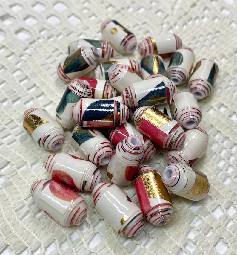 May include: A collection of cylindrical beads featuring a white base with layered paper patterns in shades of red, teal, and metallic gold. Each bead has a central hole and a smooth, glossy finish, showcasing intricate rolled paper textures.