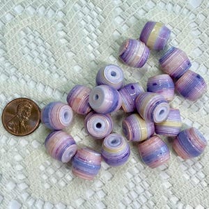 K&ouml;nnte beinhalten: Eine Ansammlung zylindrischer Perlen mit horizontalen Streifen in Lavendel, Violett, Creme und zartem Rosa. Jede Perle hat ein mittiges Loch. Eine kupferfarbene US-Ein-Cent-M&uuml;nze liegt zum Gr&ouml;&szlig;envergleich neben den Perlen.