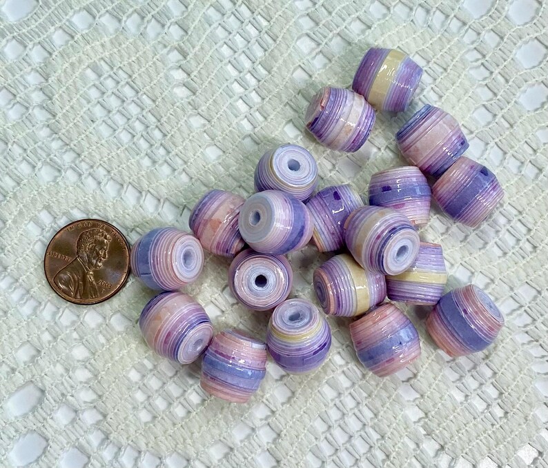 K&ouml;nnte beinhalten: Eine Ansammlung zylindrischer Perlen mit horizontalen Streifen in Lavendel, Violett, Creme und zartem Rosa. Jede Perle hat ein mittiges Loch. Eine kupferfarbene US-Ein-Cent-M&uuml;nze liegt zum Gr&ouml;&szlig;envergleich neben den Perlen.