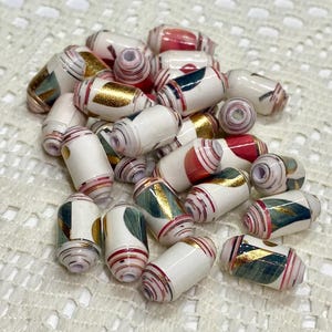 May include: A collection of cylindrical paper beads featuring a white base with decorative accents in red, teal, and metallic gold. Each bead has a central hole and is constructed from tightly rolled paper strips with visible layered edges.