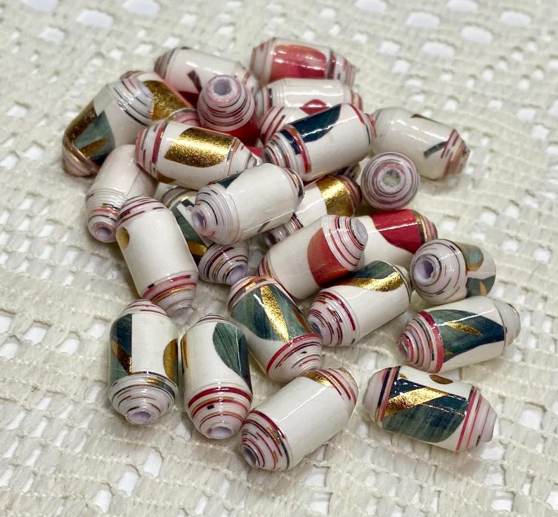 May include: A collection of cylindrical paper beads featuring a white base with decorative accents in red, teal, and metallic gold. Each bead has a central hole and is constructed from tightly rolled paper strips with visible layered edges.