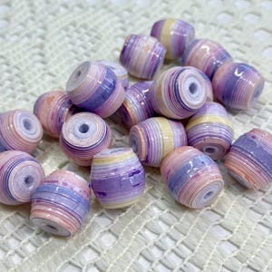 K&ouml;nnte beinhalten: Eine Ansammlung zylindrischer Perlen mit einem geschichteten, gerollten Papierdesign. Jede Perle zeigt einen Verlauf aus Pastellfarben wie zartem Rosa, Lavendel und Creme, mit einer gl&auml;nzenden Oberfl&auml;che und einem zentralen Loch zum Auff&auml;deln.