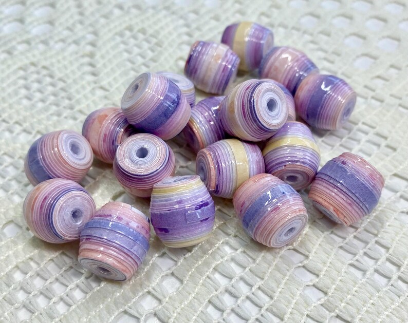 K&ouml;nnte beinhalten: Eine Ansammlung zylindrischer Perlen mit einem geschichteten, gerollten Papierdesign. Jede Perle zeigt einen Verlauf aus Pastellfarben wie zartem Rosa, Lavendel und Creme, mit einer gl&auml;nzenden Oberfl&auml;che und einem zentralen Loch zum Auff&auml;deln.