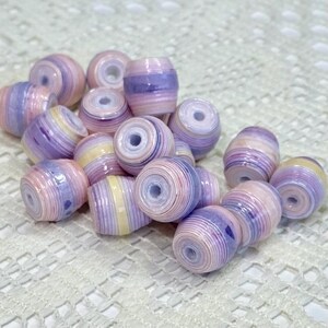 K&ouml;nnte beinhalten: Eine Ansammlung zylindrischer Perlen mit einem geschichteten, gerollten Papierdesign in sanften Pastellt&ouml;nen wie Lavendel, Hellrosa und Creme. Jede Perle hat ein mittiges Loch und eine glatte, gl&auml;nzende Oberfl&auml;che mit konzentrischen Farbringen.