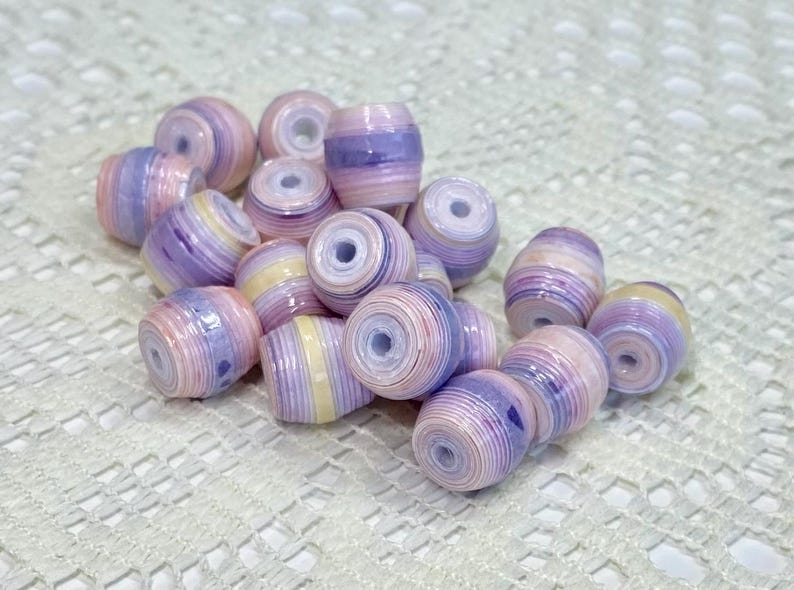 K&ouml;nnte beinhalten: Eine Ansammlung zylindrischer Perlen mit einem geschichteten, gerollten Papierdesign in sanften Pastellt&ouml;nen wie Lavendel, Hellrosa und Creme. Jede Perle hat ein mittiges Loch und eine glatte, gl&auml;nzende Oberfl&auml;che mit konzentrischen Farbringen.