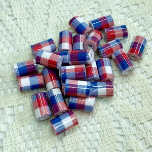 May include: A collection of small, cylindrical beads with a red, white, and blue plaid pattern. The beads are scattered on a textured, off-white surface. The plaid design is consistent across all beads.