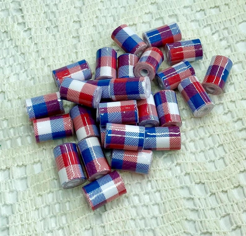 May include: A collection of small, cylindrical beads with a red, white, and blue plaid pattern. The beads are scattered on a textured, off-white surface. The plaid design is consistent across all beads.