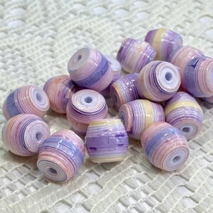 K&ouml;nnte beinhalten: Eine Ansammlung von tonnenf&ouml;rmigen Perlen mit einem geschichteten, gerollten Papierdesign. Jede Perle zeigt eine Farbpalette aus zarten pastellfarbenen violetten, hellrosa, cremefarbenen und wei&szlig;en Streifen mit einem mittigen Loch.
